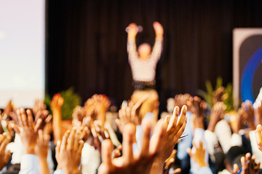 blurred photograph of a person on stage with their arms raised up and a full audience of people with also their hands in the air, imitating the speaker