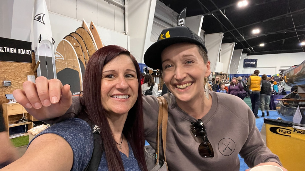 A selfie taken by Christina, a woman with long dark red hair. Tarzan has her arm around Christina. She wears a black baseball cap and sunglasses hooked around the neck of her sweater. In the background you see many people, canoes, and paddleboards, and you can see they are at a massive trade show.