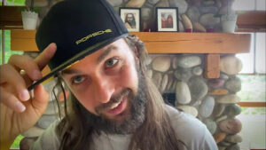 A white man appearing to be in his thirties with a messy beard and long stringy brown hair, nods at the camera and tips his black baseball cap with a neon Porsche logo. In the background is a large stone fireplace with several framed pictures of Indian men on the mantle.