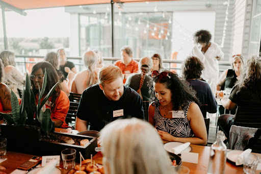 A diverse group of adults engaged in conversation at a networking event. The setting is a lively, well-lit indoor venue with modern decor. In the foreground, a man with blond hair is conversing with a woman with shoulder-length black hair, both are smiling and looking at a document one of them is holding. Other attendees are in the background, sitting or standing while talking and enjoying the event. Everyone is casually dressed, suggesting a relaxed and friendly atmosphere.