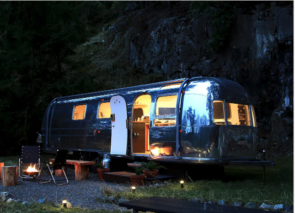 Tucked into a remote, natural environment, a polished, silver Airstream trailer is parked on a patch of grass and gravel, with soft warm light glowing from inside, illuminating its interior through several windows. The trailer door is partially open, revealing hints of a cozy, well-lit living space. Outside, the campsite features a small setup: two foldable black chairs positioned around a metal firepit with a glowing fire, several wooden benches, and a few potted plants placed on wooden platforms. Small ground lights are scattered along the edge of the gravel area, adding ambiance to the outdoor space. In the background, a rock face rises up, partially covered in foliage.