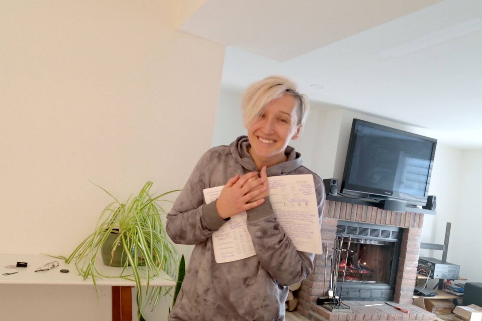 Tarzan is smiling while holding an open journal close to her chest, jammed with handwritten notes all over it. She wears a gray, tie-dye Lululemon hoodie and stands in a bright room with a fireplace, a mounted TV, and a potted plant on a nearby surface. 