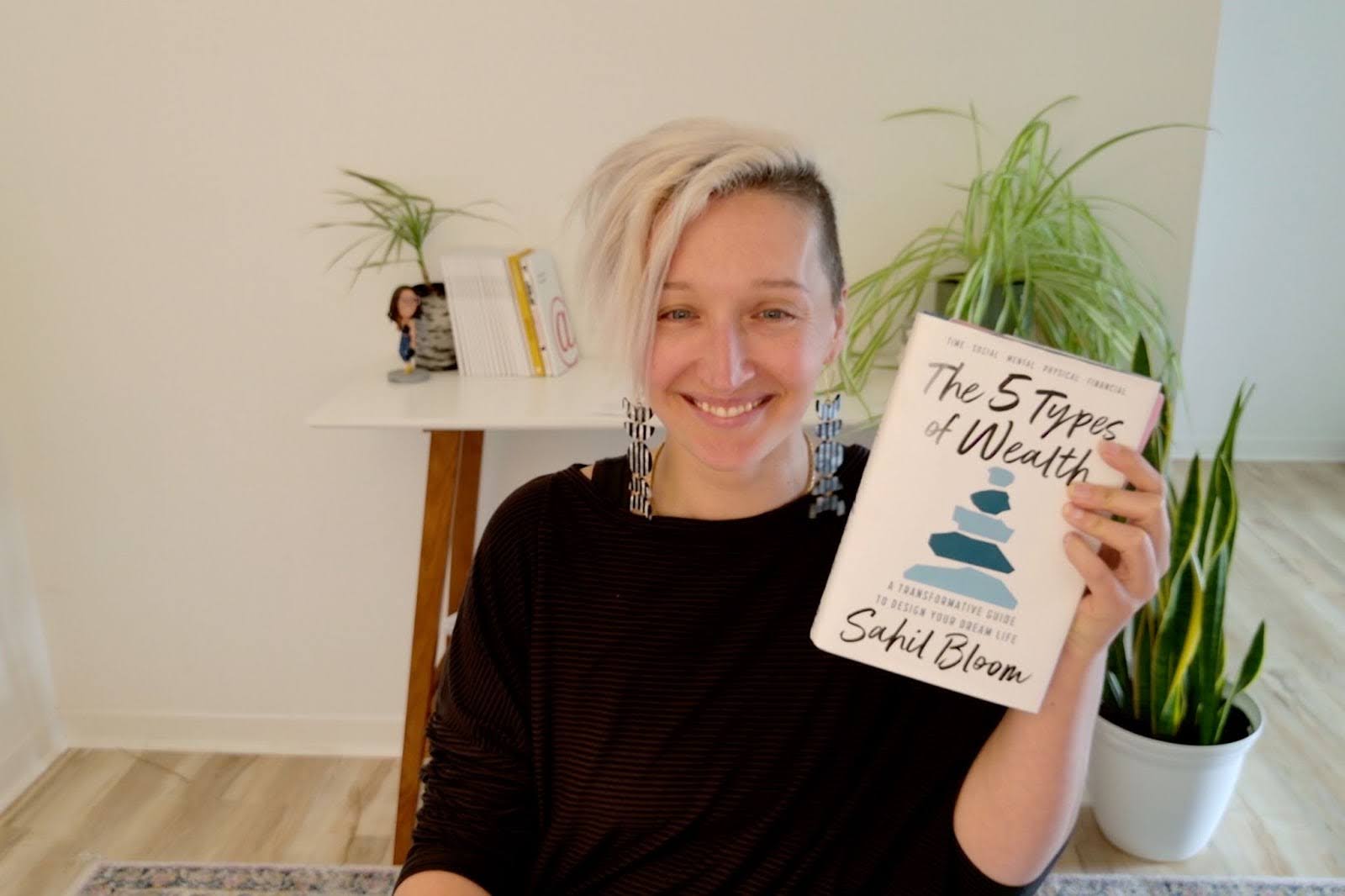 Tarzan sits at her desk, hair short, asymmetrical platinum blonde, wearing long geometric earrings and smiling. They are holding up a book titled The 5 Types of Wealth by Sahil Bloom. Behind them is a white wall, a wooden desk with books and small plants, and a potted snake plant on the floor. The space is bright and minimalistic.