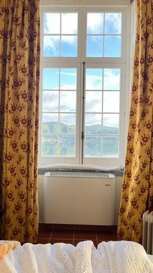 A bright, sunlit window with white frames and multiple glass panes, looking out onto a scenic landscape of green hills under a partly cloudy blue sky. The window is flanked by yellow curtains with a red floral pattern. Below the window is a white radiator or heater. In the foreground, a white textured blanket and a towel are partially visible on a bed.