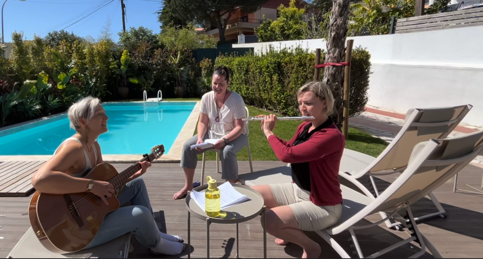 Three people are gathered on a wooden deck beside a swimming pool on a sunny day. One person sits on a lounge chair playing an acoustic guitar, another person is playing a silver flute while seated, and a third person sits on a white stool holding a notebook and smiling. There are tropical plants and hedges surrounding the pool, and a few modern buildings are visible in the background. A small round table with sheet music and a yellow water bottle is in the foreground.