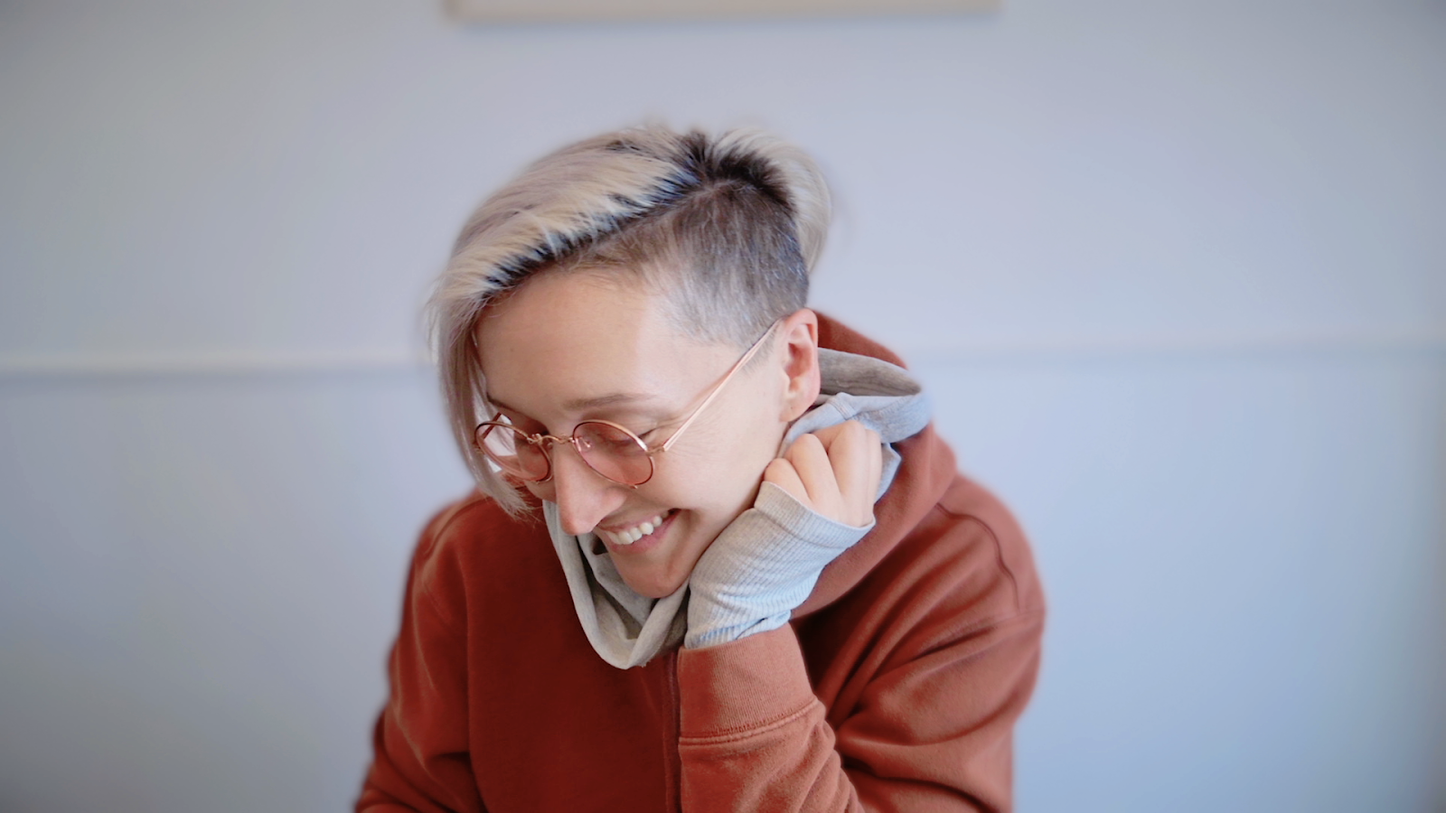 Tarzan is smiling and looking down, her hair is  partially shaved, light blonde on top and dark underneath . She's wearing round, rose-tinted glasses and a rust-coloured hoodie with light grey sleeve cuffs. One hand is touching the side of her face in a gesture that suggests shyness or laughter. The background is softly blurred in neutral tones.