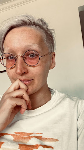 Tarzan, with short, platinum blonde hair and light skin is shown in a close-up selfie. She is wearing round, wire-rimmed glasses with a rose tint and a white shirt featuring an abstract orange design. The individual is resting their chin on their hand and looking directly at the camera with a thoughtful expression. The background is an indoor setting with off-white walls and part of a doorframe visible.