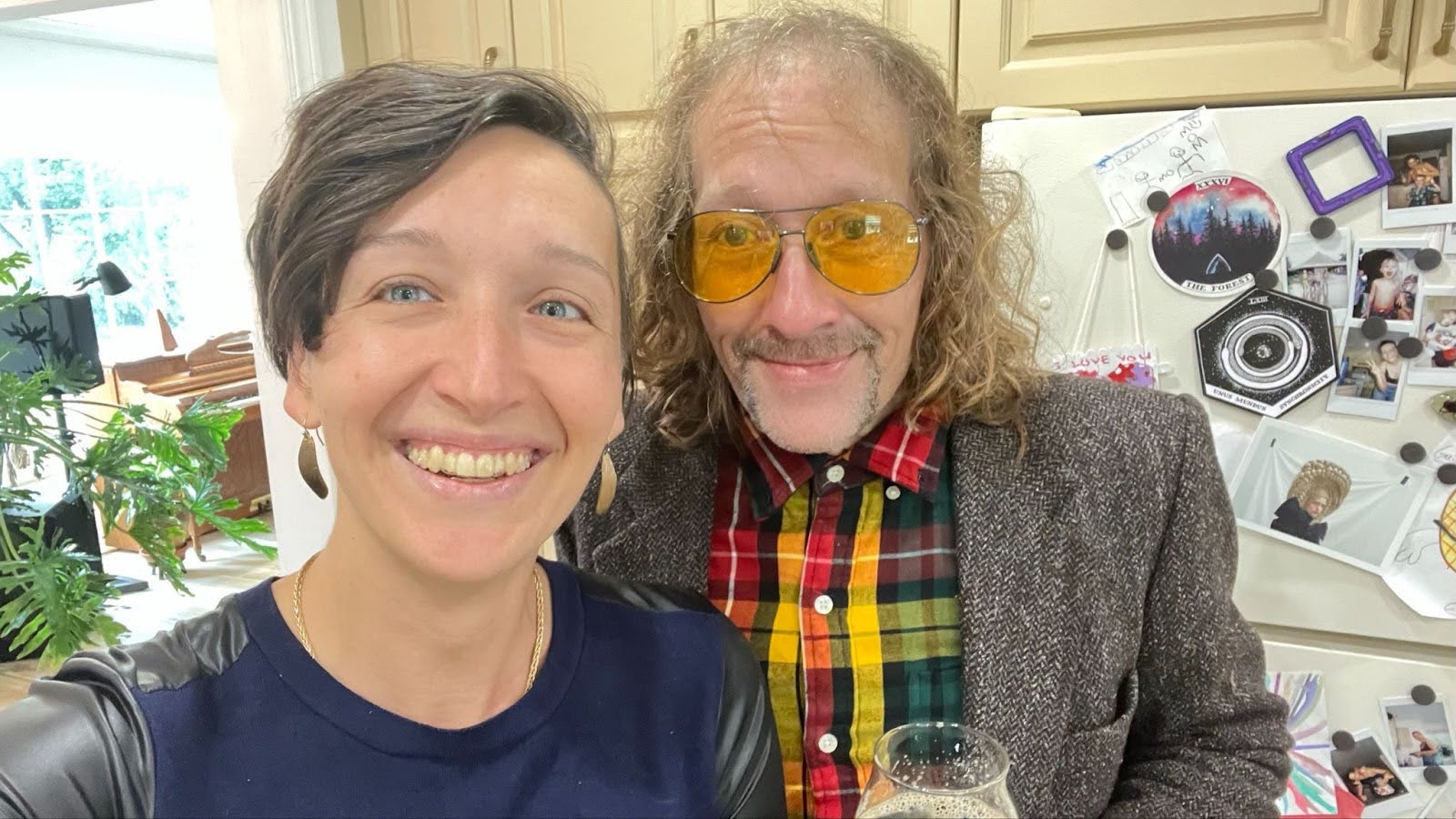 A selfie of Tarzan and her brother Quentin, both smiling at the camera. Tarzan wears a navy blue top with leather sleeves, small earrings, and her short brown hair sweeping down one side of her face; she has a wide, open smile and blue eyes. Quentin wears orange-hued aviator sunglasses, a plaid shirt with a sports jacket, and holds a beverage. He has shoulder-length wavy hair, a mustache, and a chin strap beard, with a subtle yet radiant smile. Behind them is a fridge with family photos, notes, and stickers. A plant, a piano, and bright window are visible in the background.
