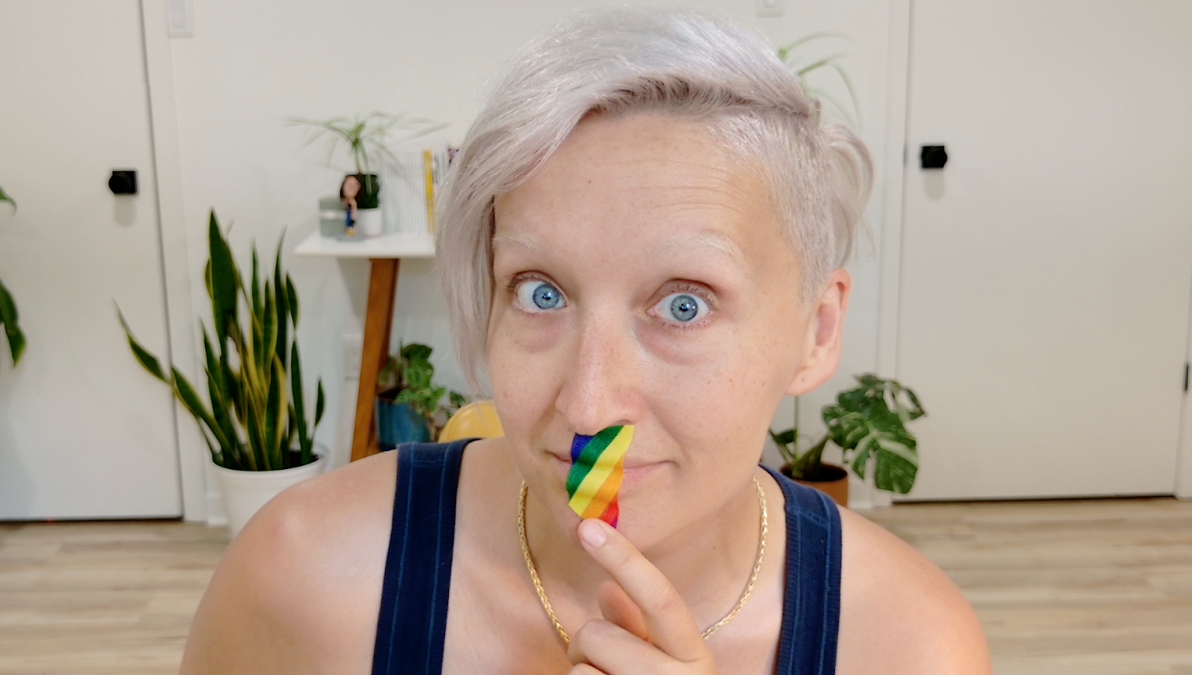 Tarzan lips are taped with rainbow-striped hockey tape. She has light-coloured hair, bright blue eyes and is wearing a dark sleeveless top and a gold chain necklace. The background shows a room with white walls, potted plants, a small shelf with books and a figurine, and wooden flooring. She's looking directly into the camera with a playful expression.