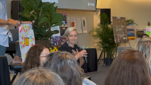 Tarzan engaging with audience during a collaborative workshop, holding a microphone while seated near visual boards covered in colorful sticky notes, surrounded by plants and event materials.