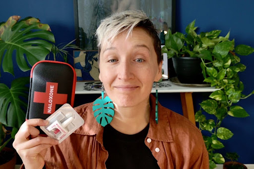 Tarzan, with short, two-tone hair sits indoors in front of leafy houseplants, holding up a black naloxone kit and a nasal spray device. She's wearing large teal leaf-shaped earrings and a brown shirt, and is looking toward the camera with a neutral expression.