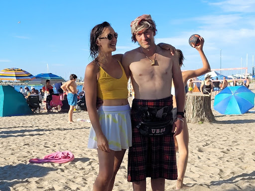 Tarzan wearing a yellow top and white skirt stands on a sandy beach with their arm around Quentin wearing a plaid kilt, a head wrap, and a cross-body pouch. They are both smiling. Behind them are beach umbrellas, people sitting in chairs, and others walking or playing sports in bright sunlight.