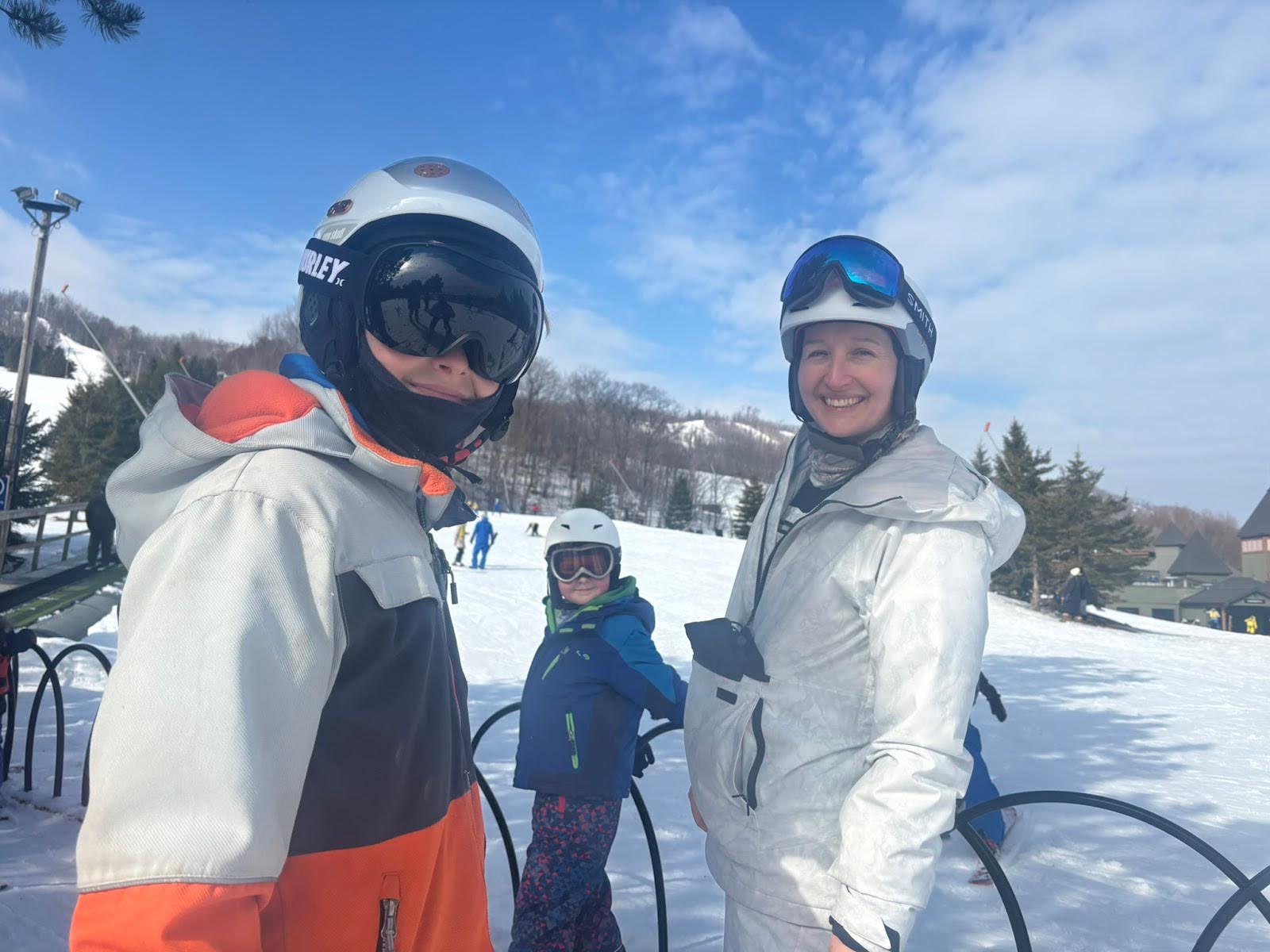Tarzan and her son in ski helmets and goggles smile at the camera on a snowy ski slope; one wears a gray-and-orange jacket and the other a white snowsuit. Her youngest son in a blue jacket and helmet stands behind them near a metal railing, with snow-covered hills, trees, and a partly cloudy blue sky in the background.