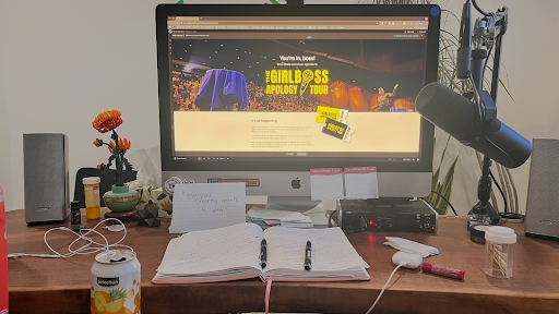 Tarzan's Home office desk setup with an iMac displaying The Girlboss Apology Tour website, surrounded by a notebook, sticky notes, microphone, speakers, coffee cup, and workspace essentials, showing behind-the-scenes marketing and planning work.