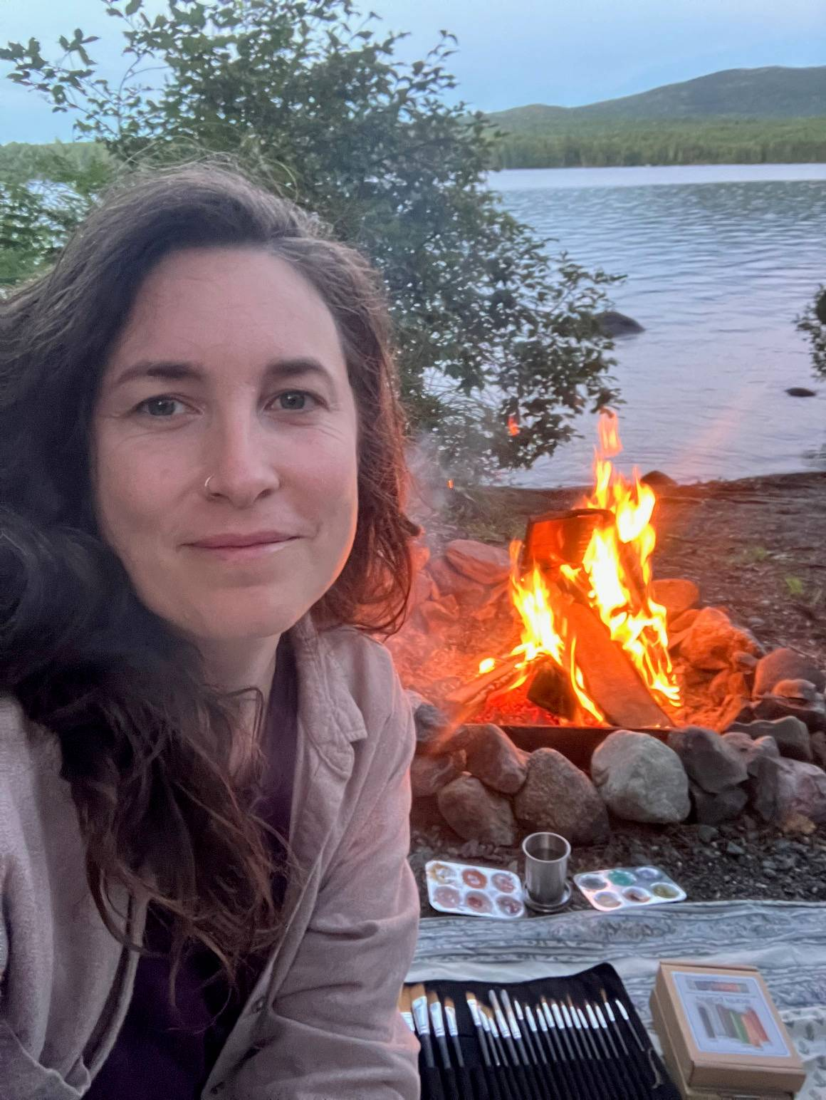 Kendra sitting by a lakeside campfire at dusk, smiling at the camera with mountains and water in the background, surrounded by art supplies and painting tools, creating a peaceful outdoor creative scene.