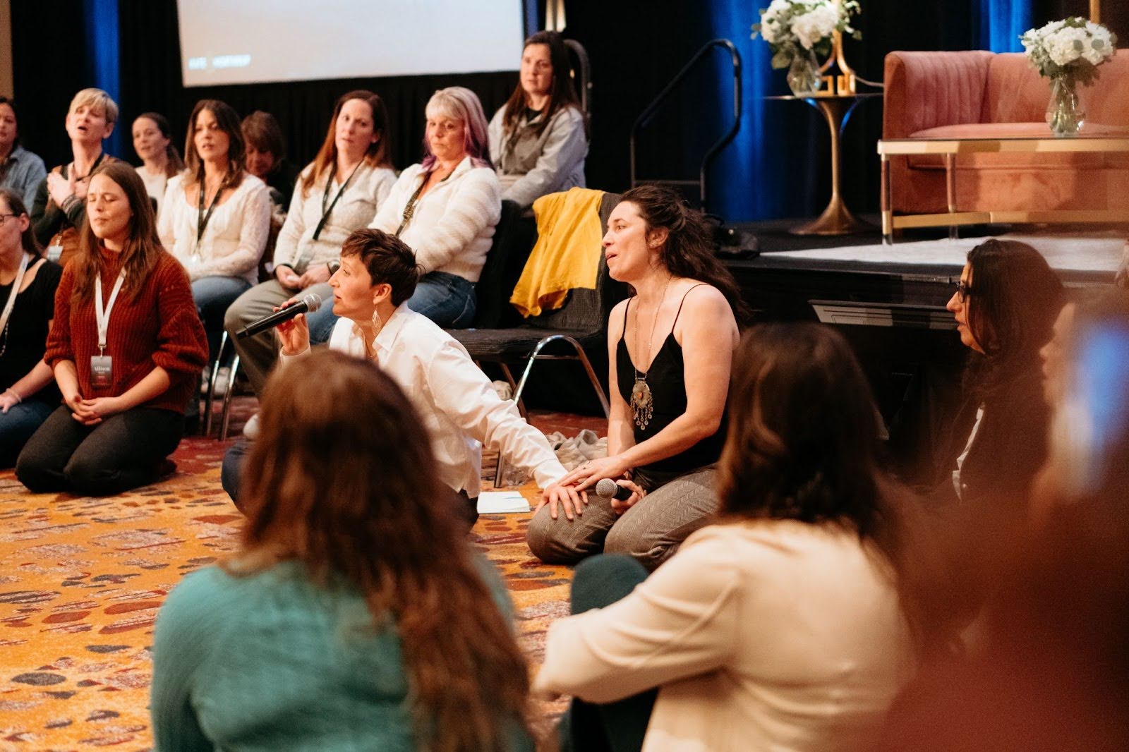 Group of women gathered in a circle at a conference, Tarzan holding a microphone and speaking or singing during an interactive workshop session, while others listen and engage in a collaborative, community-focused environment.