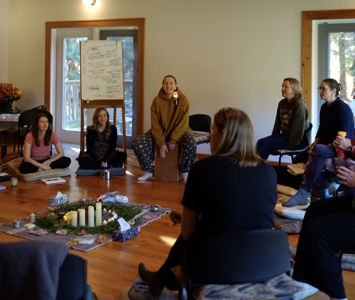 A group of women sit in a circle on cushions and chairs in a warm, sunlit wooden room, singing In the center, a centerpiece of evergreen boughs, lit candles, and small objects is laid out on a woven rug. Tarzan is smiling, in a mustard hoodie, and sits on a wooden box drum beside a flip chart covered in handwritten notes, while the others listen and sing.